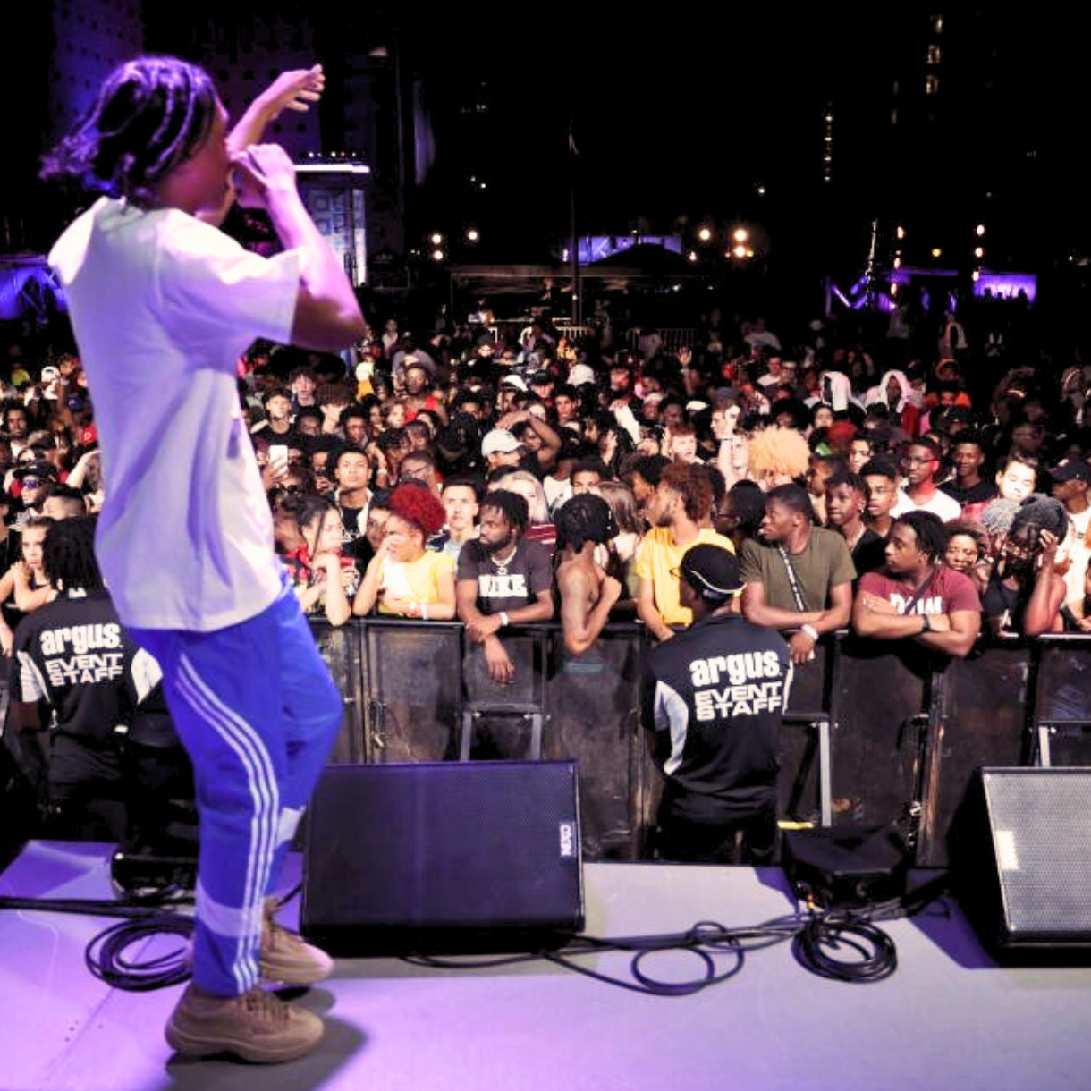 A performer stands on stage holding a microphone, engaging a large crowd at a nighttime outdoor music event.