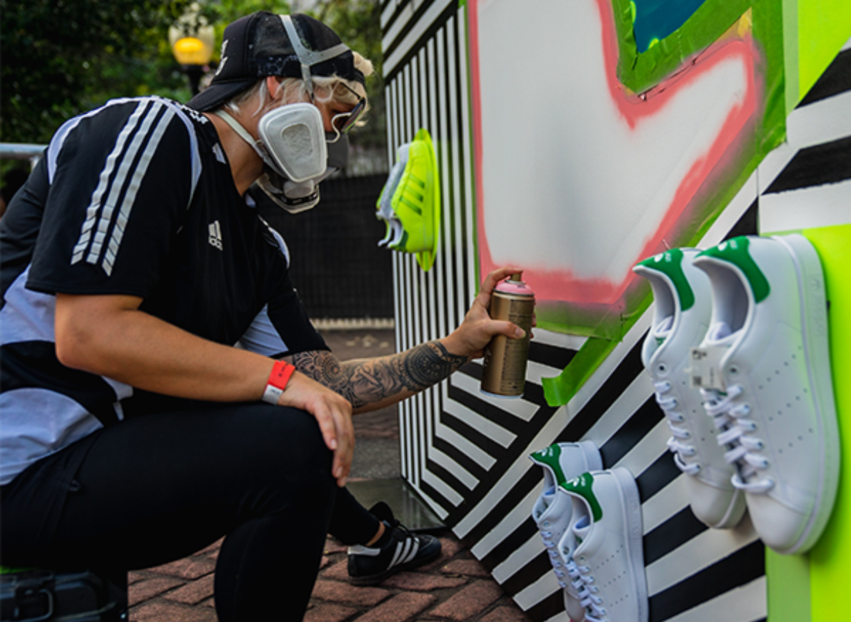 An artist wearing a mask spray-paints a graphic Adidas installation with shoes displayed on the wall.