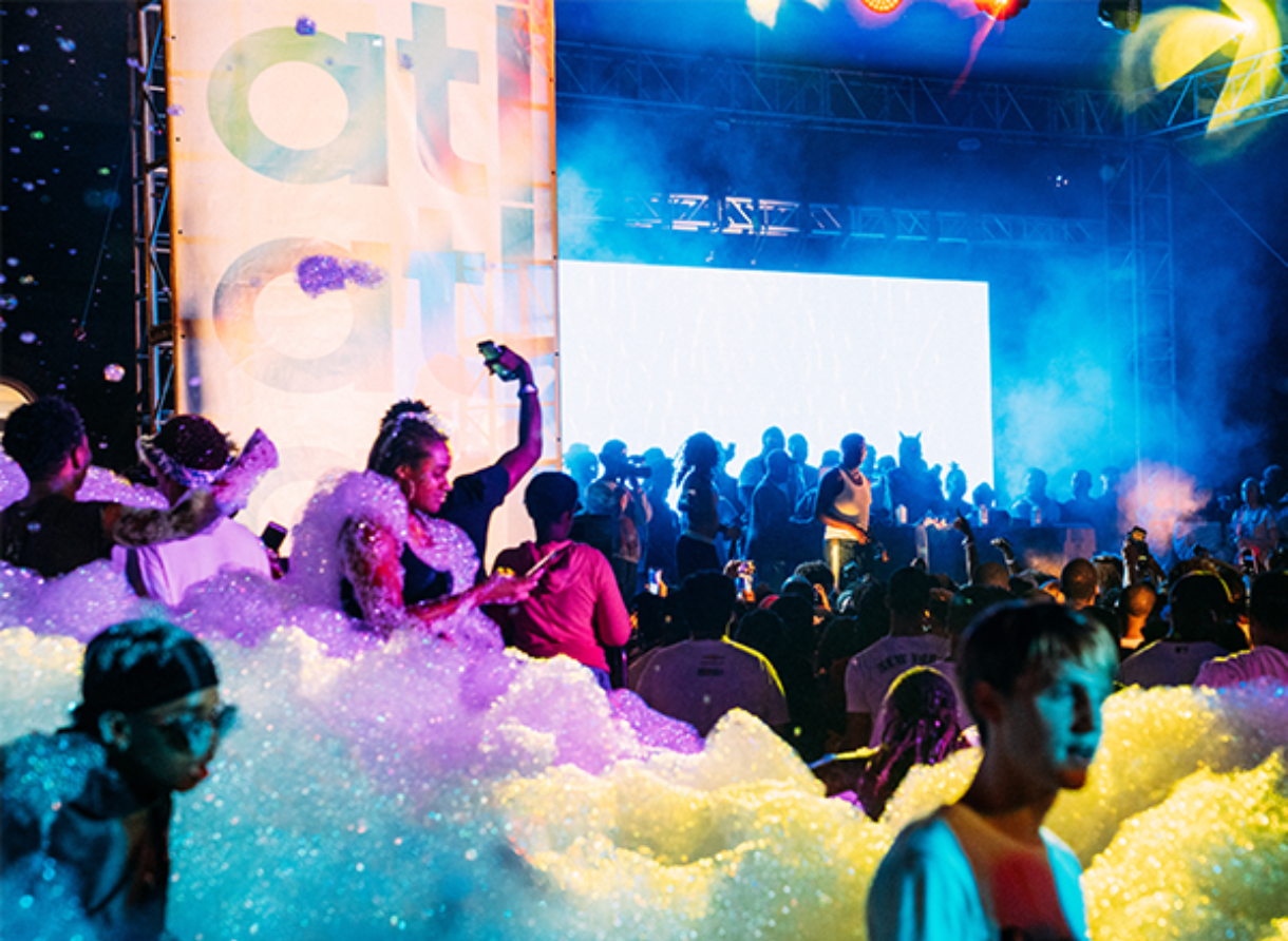A large crowd gathers at an outdoor foam-filled music event, with colorful lights and a stage in the background.
