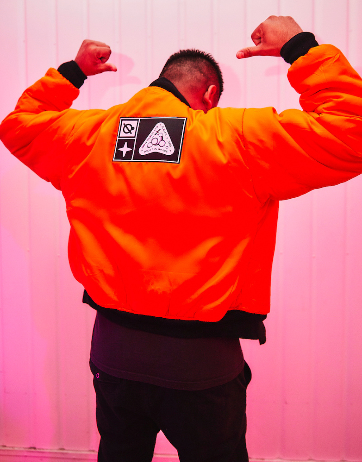 Guest wearing a bright orange jacket with custom Goodbye Gravity patches poses with arms raised against a pink-lit backdrop.