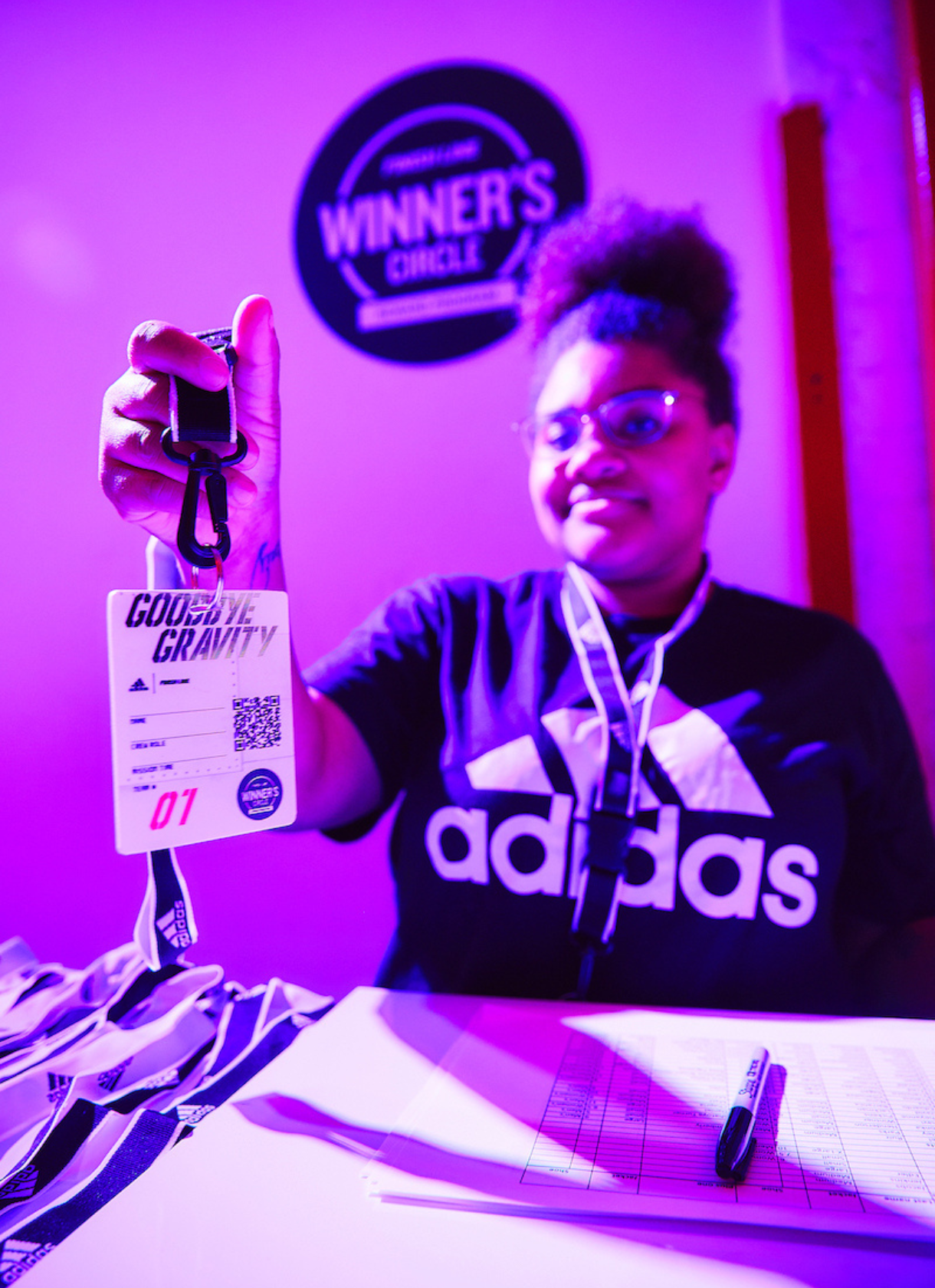 Event staff member holds up a “Goodbye Gravity” adidas event badge under purple lighting at the check-in table.