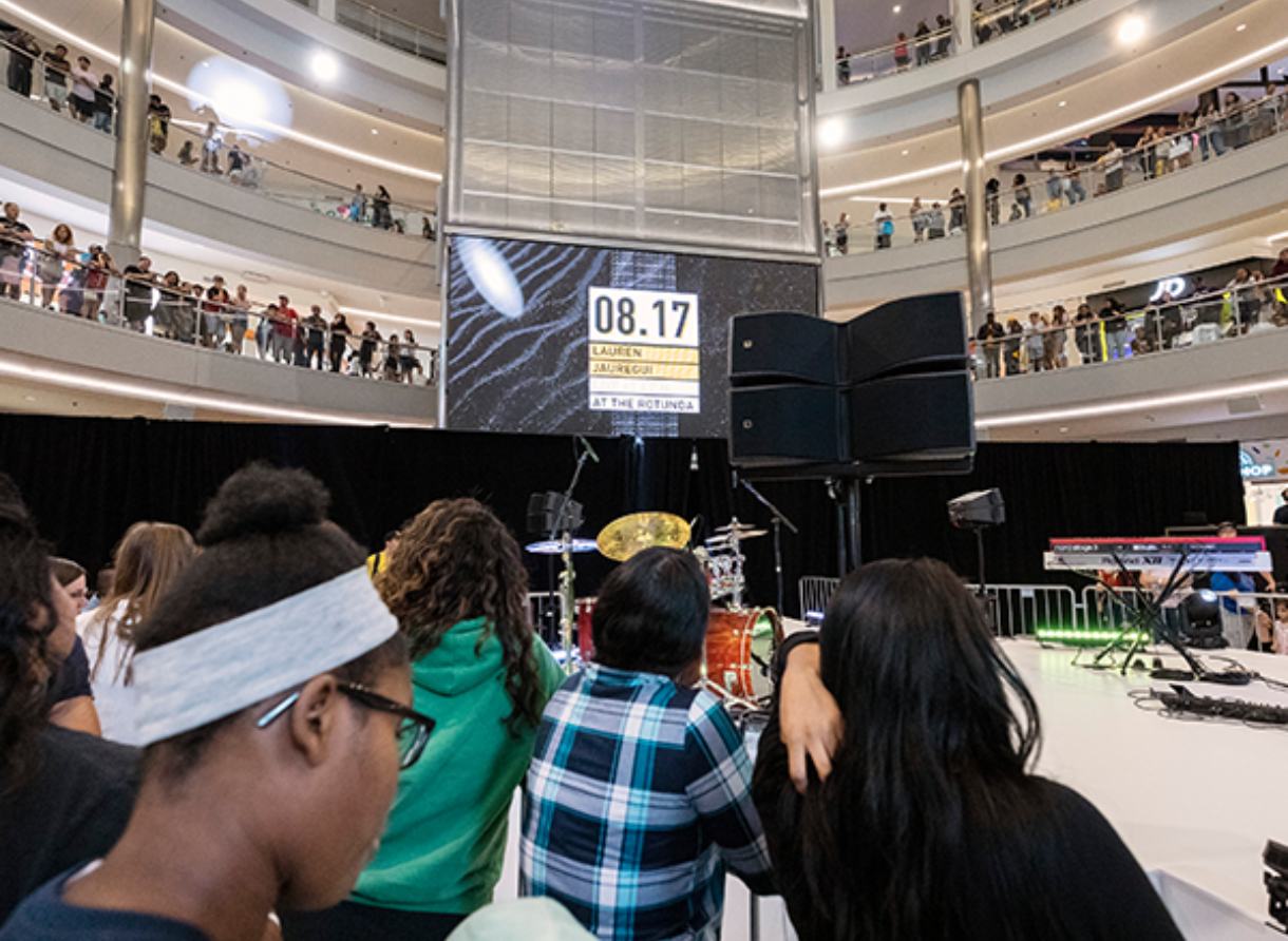 Audience watching a stage with instruments and a large digital screen displaying event information.