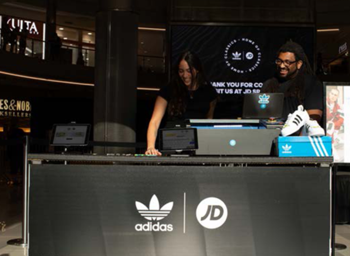 Two event staff members stand behind a custom engraving station with adidas and JD Sports branding.