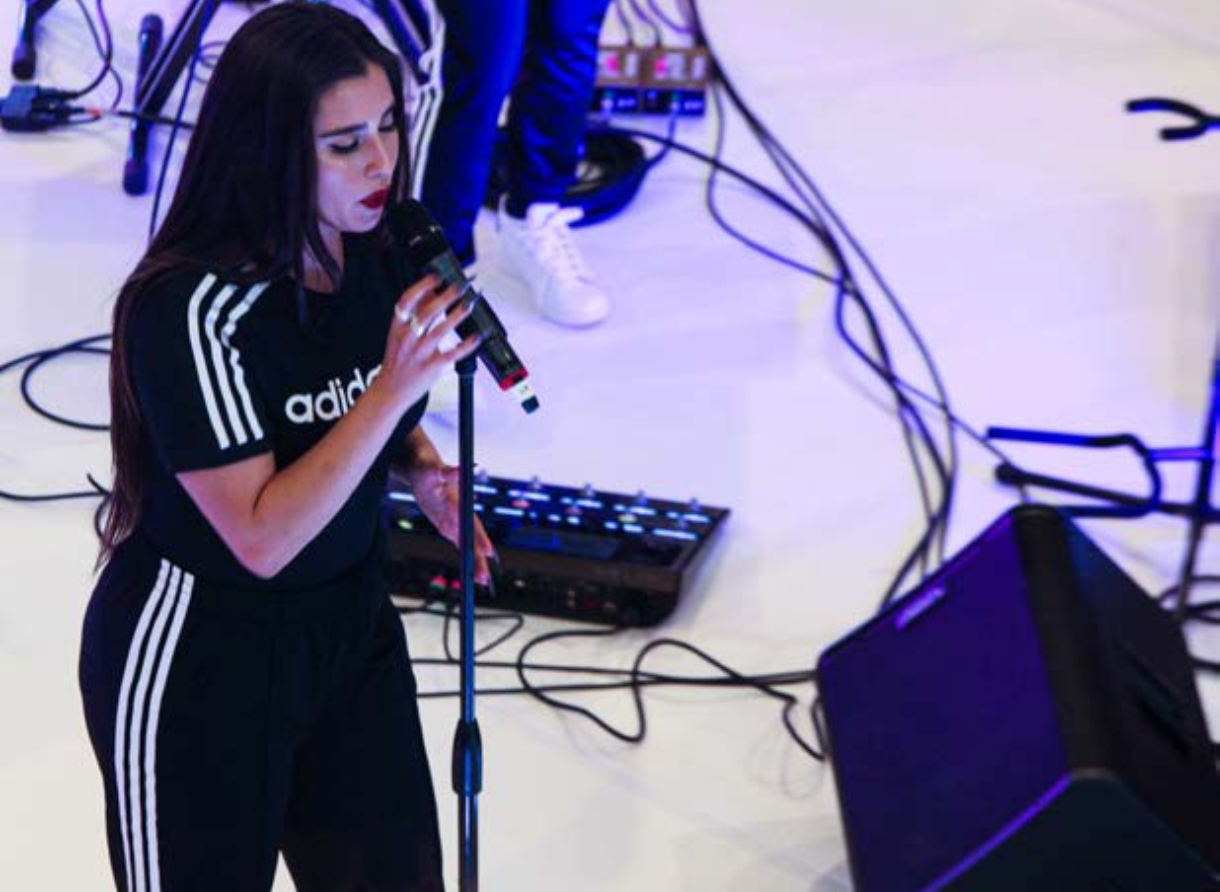 Overhead view of a live performance with an artist singing into a microphone on a white stage surrounded by equipment.
