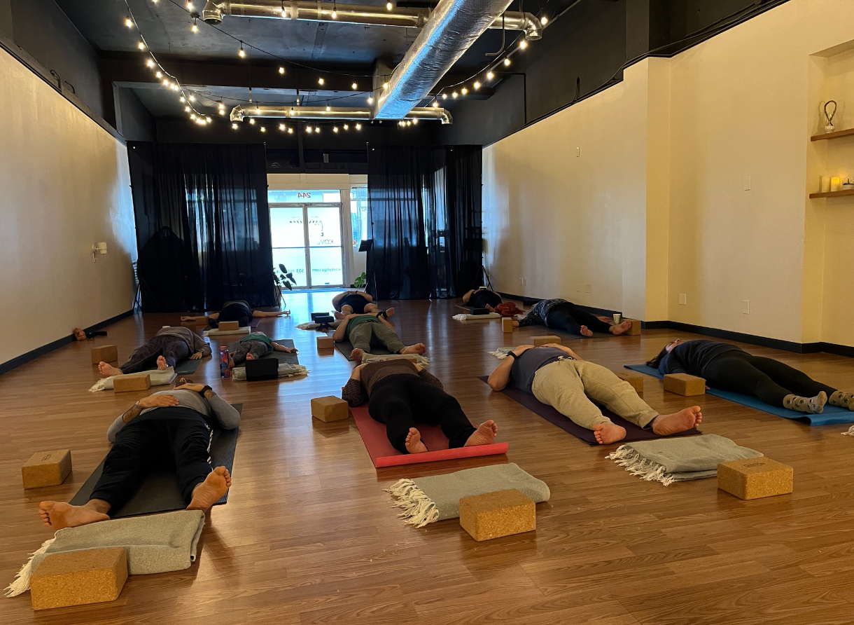 Students resting in savasana on yoga mats with bolsters and blocks in a softly lit studio.