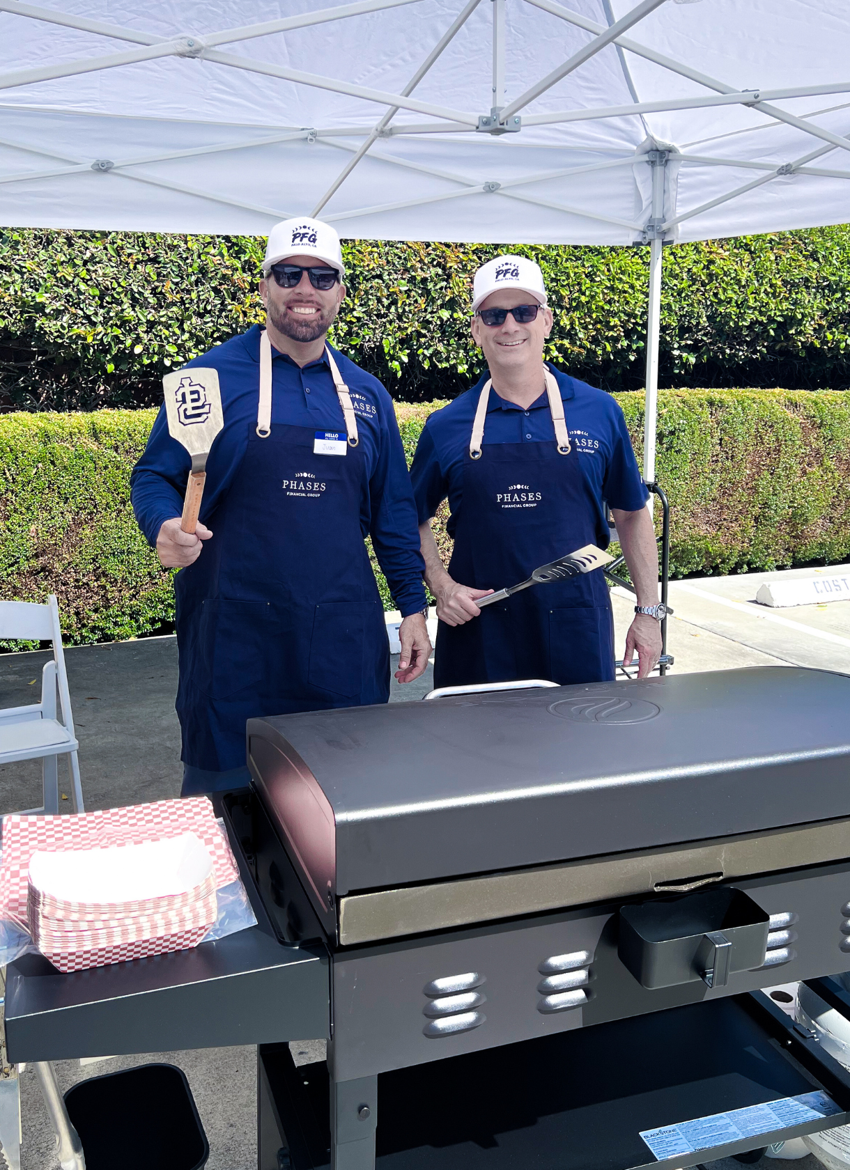 Two Phases Financial Group advisors wearing branded aprons and hats while standing behind a grill at an outdoor community event.