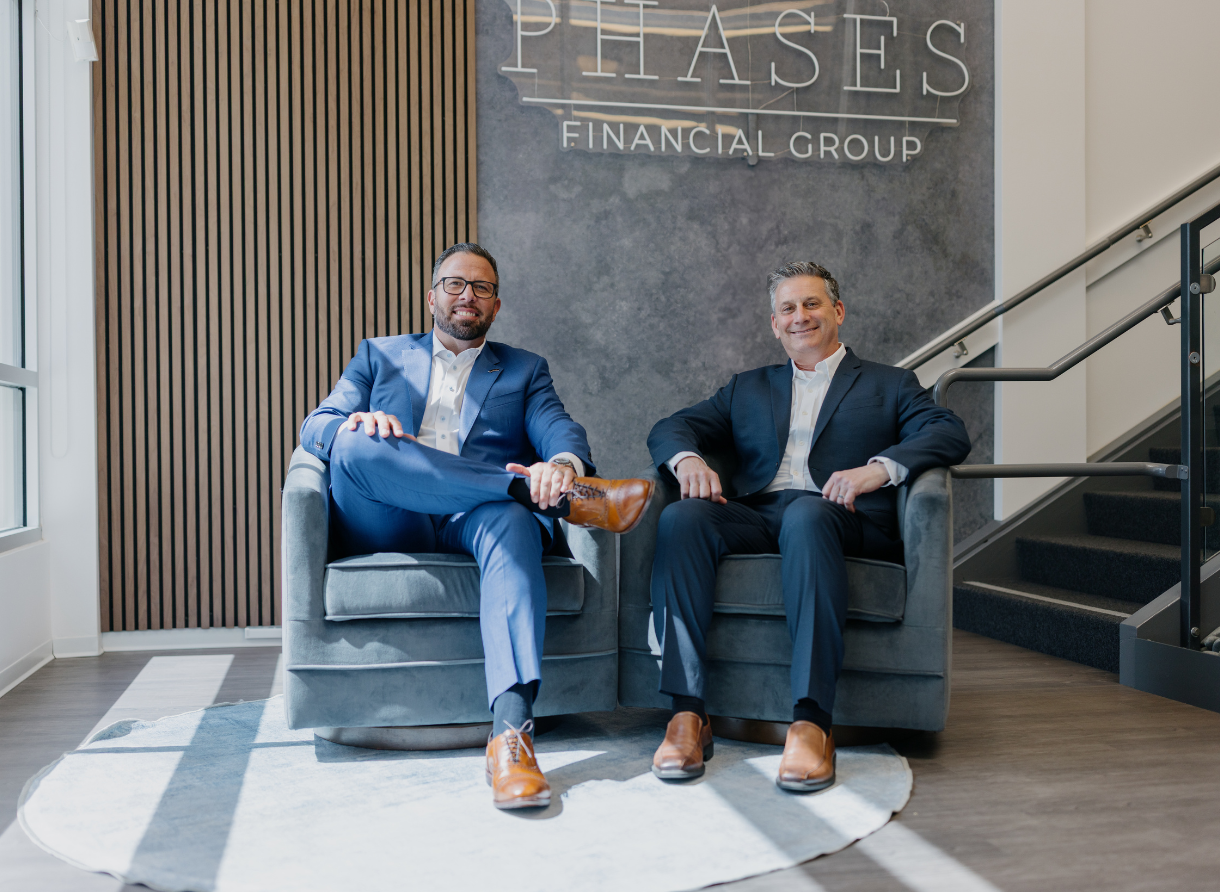 Two Phases Financial Group advisors seated in modern lounge chairs inside the Phases office, positioned beneath the illuminated Phases Financial Group logo.